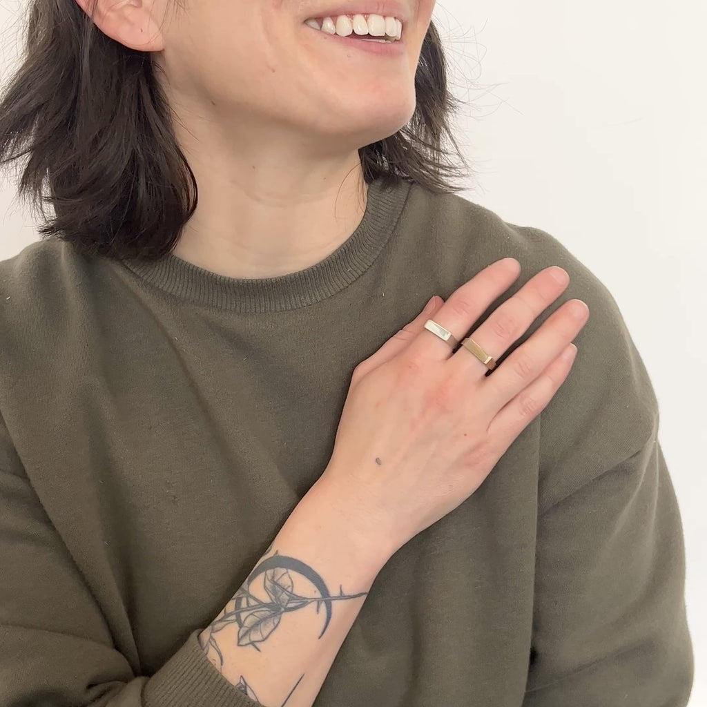 A model wears a narrow signet ring with a polished rectangular top and hammered detail on the band. The sterling silver version is in the pointer finger and the bronze is on the middle finger. They move their hand side to side. The Baliza Ring is designed and handcrafted in Portland, Oregon.
