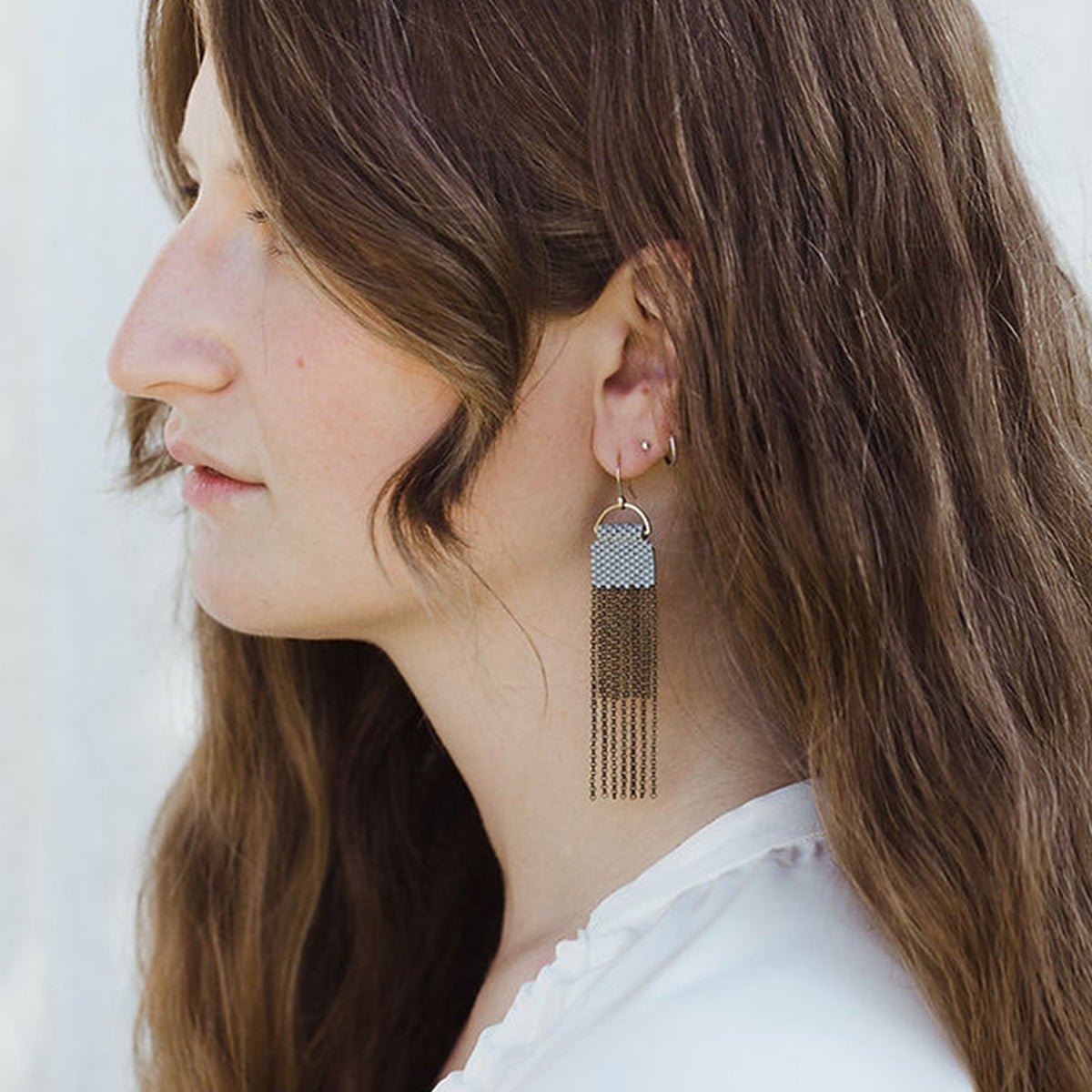 A pair of blue/grey beaded earrings with long strands of antique brass chain secured around a small brass hoop. The Curtain Earrings in Blue Grey are designed and handcrafted by A Nod to Design in Portland, OR.