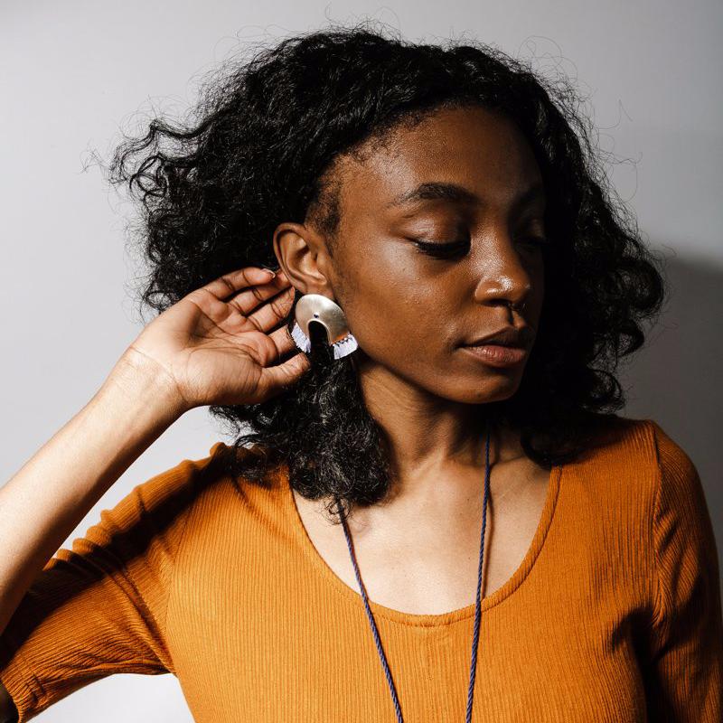 Oversized, arc-shaped bronze earrings with light blue cotton fringe and lapis lazuli; worn on a model with curly black hair and an ochre dress.