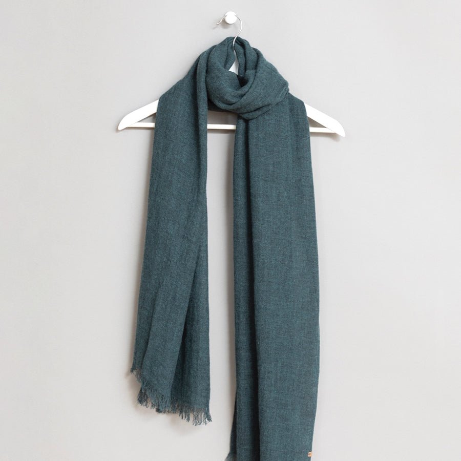 Green scarf wrapped around a white wooden hanger. The scarf has frayed edges. The Merino Woven Scarf in Dragonfly Green is from designer Dinadi.
