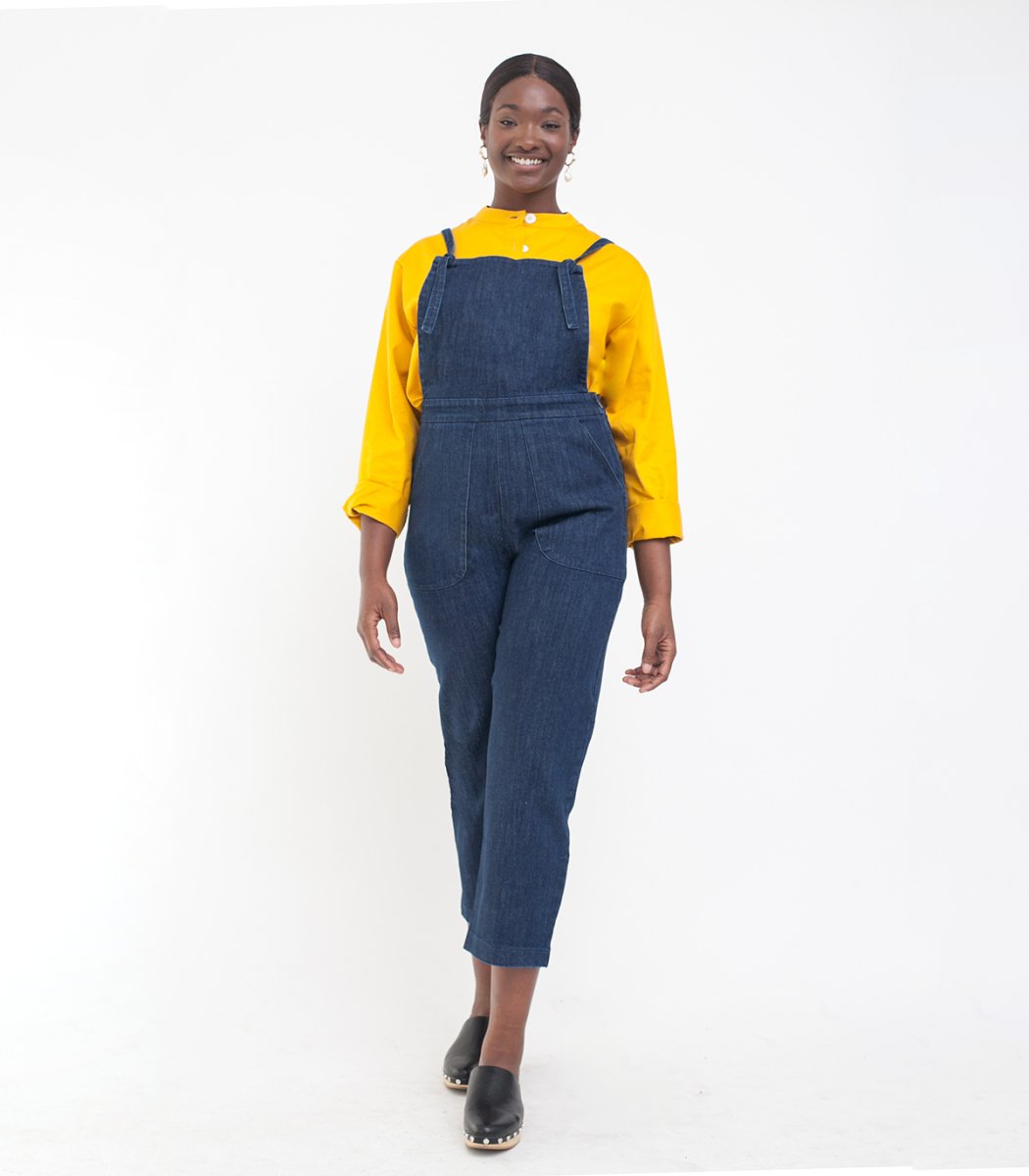 Model wears dark blue cropped overalls with thin adjustable straps and two front pockets over a long sleeve yellow shirt. The Knot Overalls in Dark Indigo are designed by Loup and made in New York City, NY.