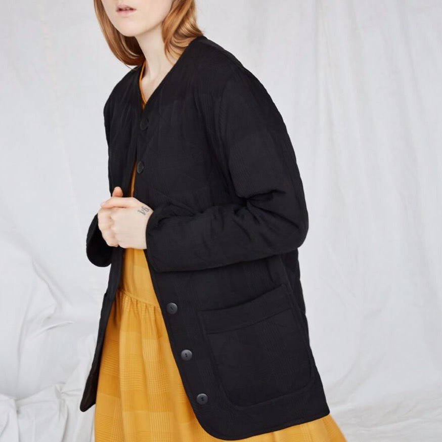 A standing red-haired woman wears a black coat and yellow dress. The Cost Jacket in Black Plaid is from Canadian designer Eve Gravel.