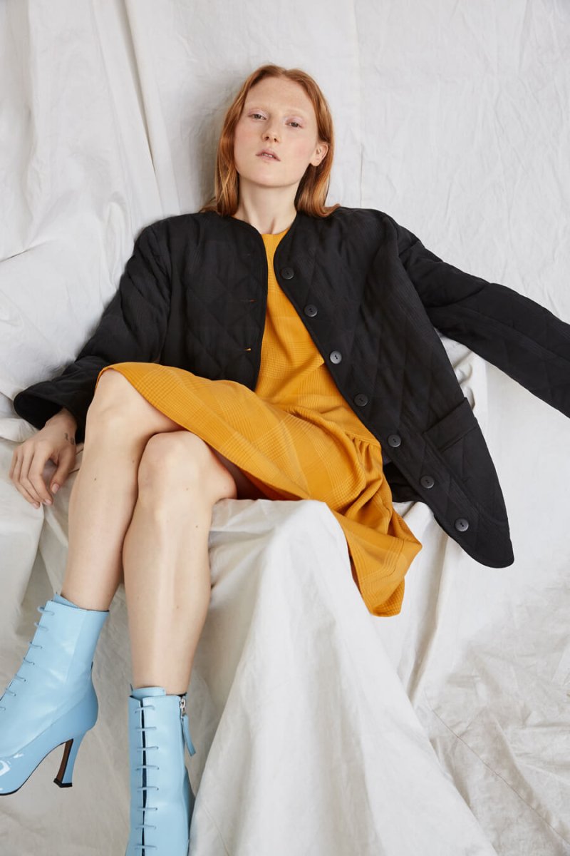 A seated red-haired woman casually wears a black coat and yellow dress. The Cost Jacket in Black Plaid is from Canadian designer Eve Gravel.