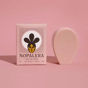 An oval shaped pink bar soap stands to the right of its pink rectangular packaging. This cactus soap contains a subtle scent of jasmine oil with rose clay. The Flor de Mayo Cactus Soap is from Nopalera and made in Brooklyn, NY.