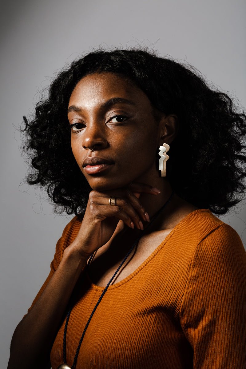 Sleek stud earrings with long curves of flat, layered bronze and silver; styled on a model with black curly hair and an ochre dress.