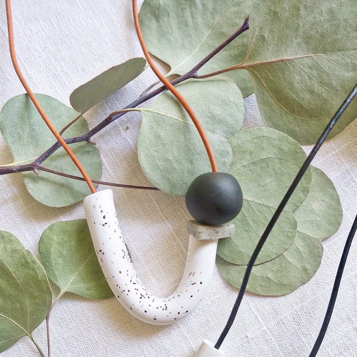 Asymmetrical white speckle U + triangle granite disc + black orbit made of polymer clay and orange leather cord. Made in Los Angeles, California by Hey Moon Designs.