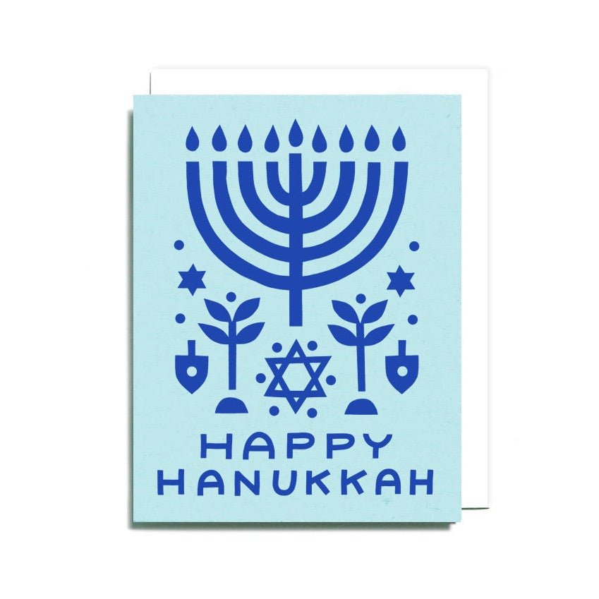 A light blue card with a dark blue festive menorah in a symmetrical paper cut collage. Front of card reads: "HAPPY HANUKKAH." Designed and handcrafted by Worthwhile Paper in Ypsilanti, MI.