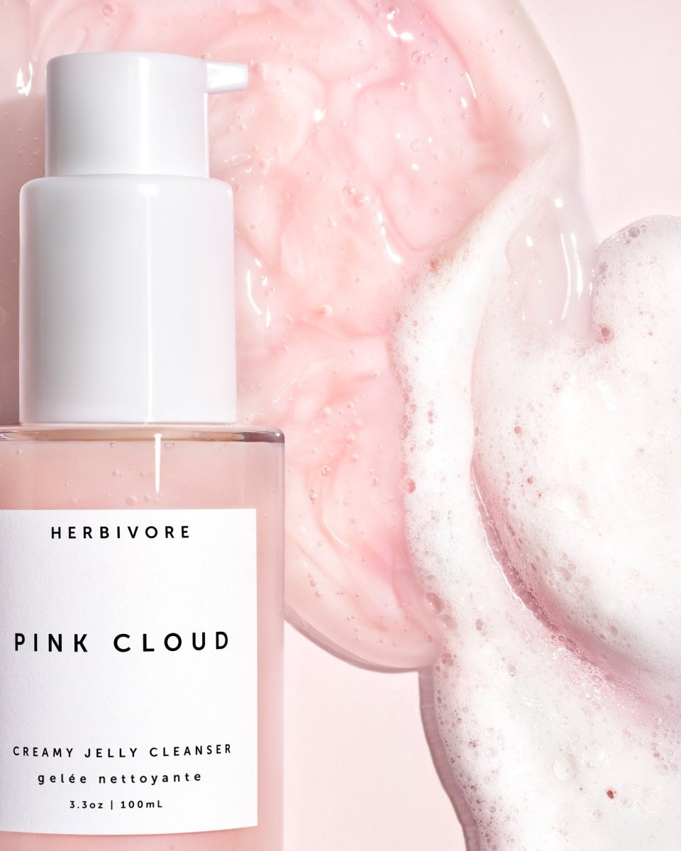 Closeup of a glass bottle with a pink foaming cleanser poured next to it. The Pink Cloud Cleanser is from the brand Herbivore.