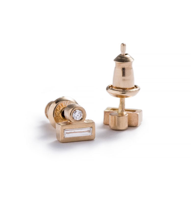 Tiny stud earrings of 14k yellow gold, featuring a small, round, white diamond set against the long side of a white diamond baguette and 14k gold earring posts and nuts. Hand-crafted in Portland, Oregon.