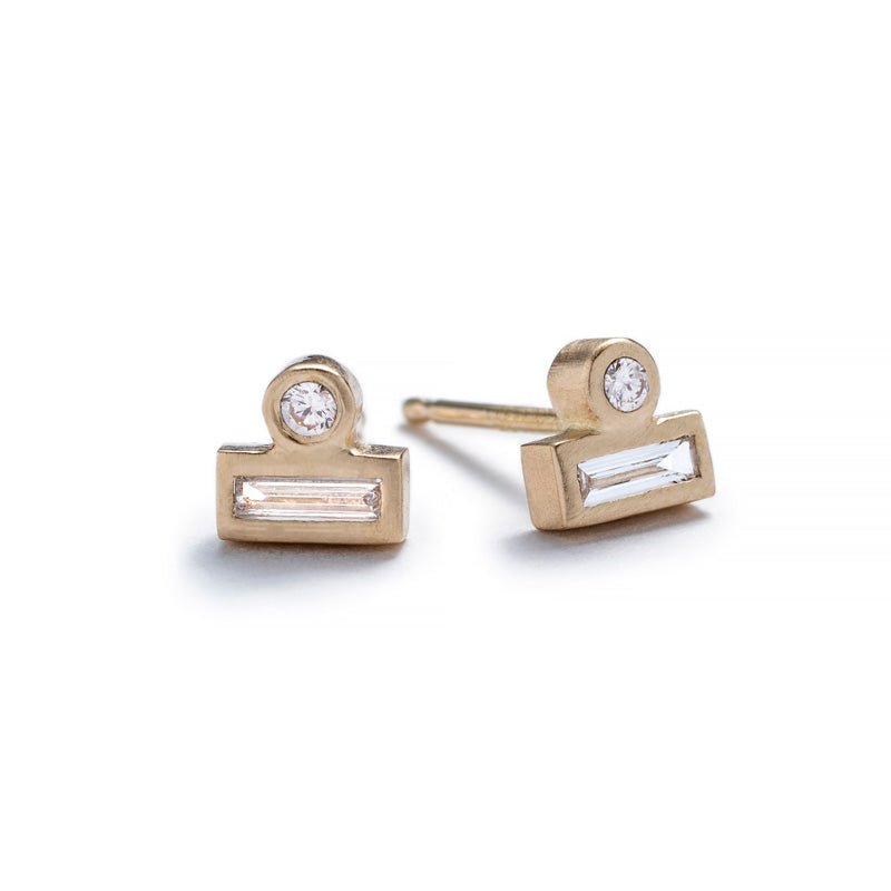Tiny stud earrings of 14k yellow gold, featuring a small, round, white diamond set against the long side of a white diamond baguette. Hand-crafted in Portland, Oregon.