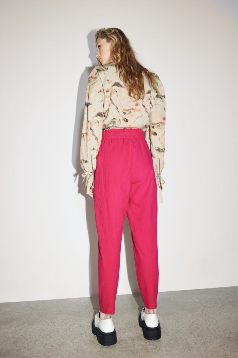 A model shows the back side of a straight leg pant in a bright pink color with an elastic waistband. The Izmir Pants in Fuchsia is designed by Eve Gravel and made in Montreal, Canada.