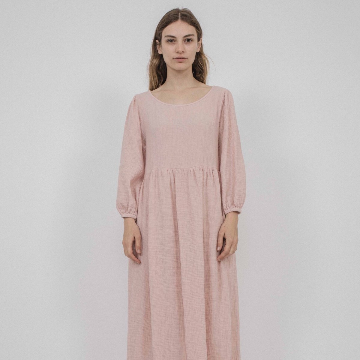 A model wears a 3/4 sleeve dress with ruching details. The dress has elastic armhole and side pockets in a light pink color. The Juniper Dress in Blush is designed by Corinne Collection in Los Angeles, CA.