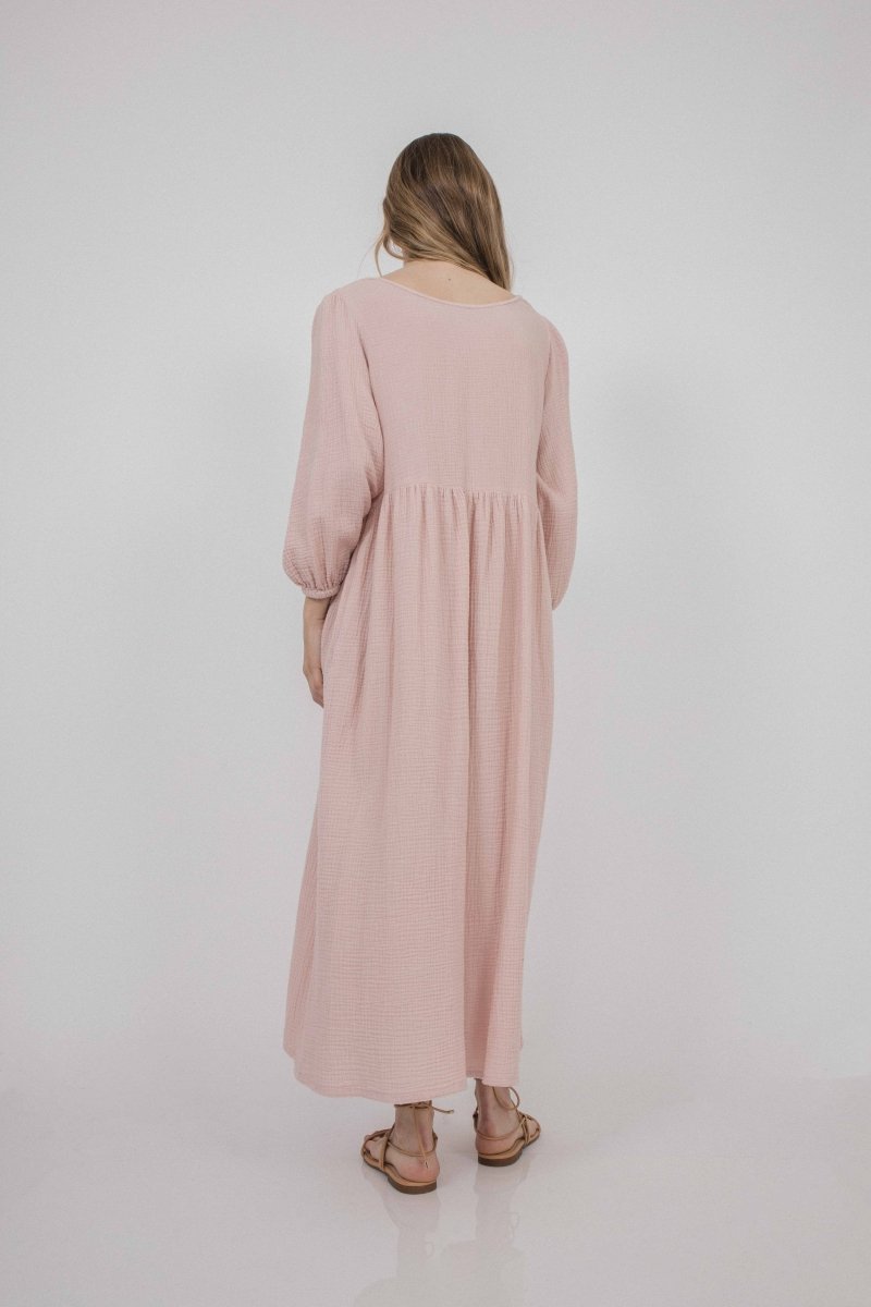 A model shows the backside of a 3/4 sleeve dress with ruching details. The dress has elastic armhole and side pockets in a light pink color. The Juniper Dress in Blush is designed by Corinne Collection in Los Angeles, CA.
