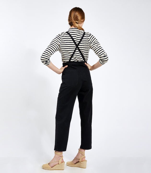 Model shows the back side of cropped black overalls with thin adjustable straps and two back pockets over a black and white striped turtleneck shirt. Overalls are backless with adjustable straps criss-crossed. The Knot Overalls in Black are designed by Loup and Made in New York City, NY.
