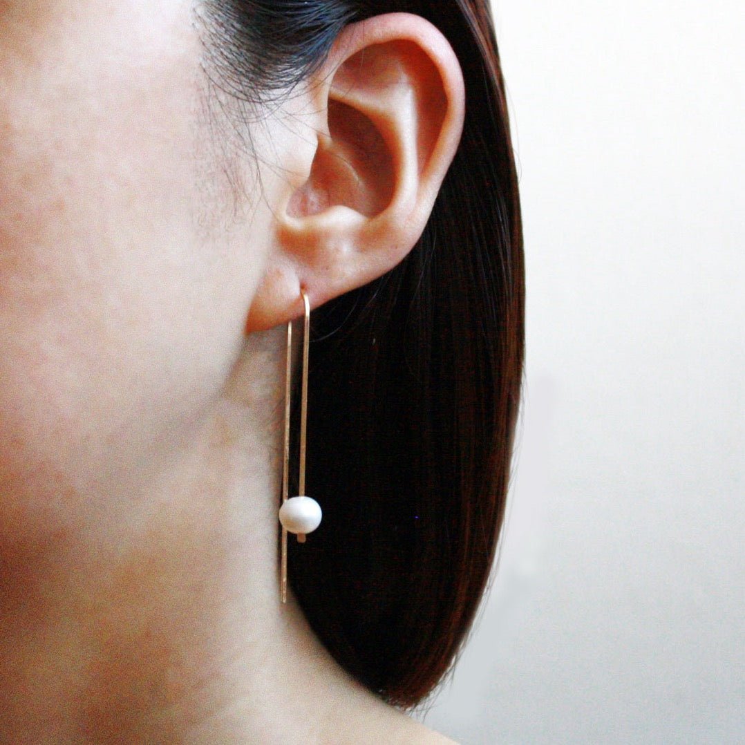 A model wears a long gold tone u-shaped earring with a single large pearl. The Long Arc Threader Earrings with Large Pearl is designed by Hooks and Luxe and handcrafted in Jackson Heights, NY.