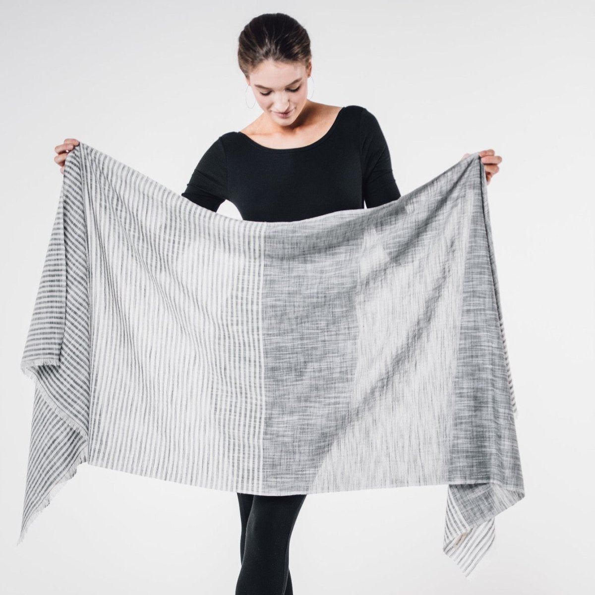 Model holds up grey scarf with white stripes and cotton fringe. The Malabar Natural Khadi Scarf in Grey is from Bloom & Give and hand-loomed in South India.