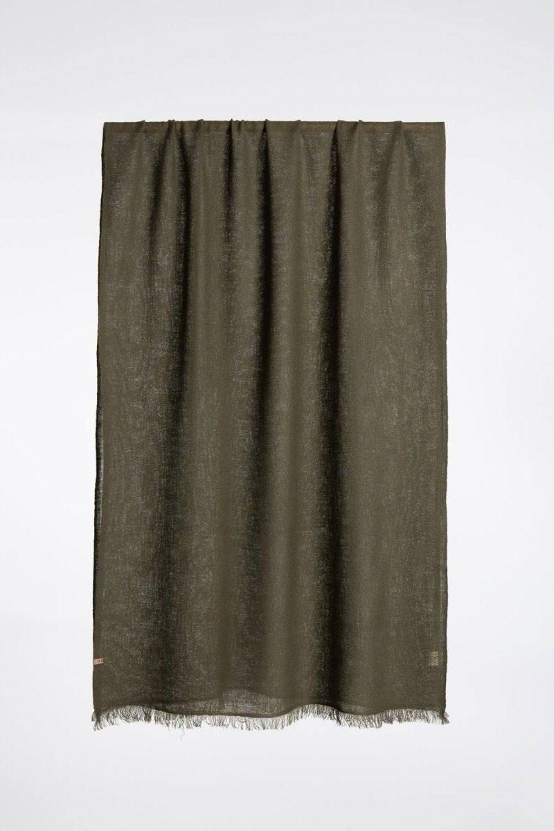 A dark green scarf made of 100% wool drapes over a wood dowel. The Merino woven Scarf in Olive Green is designed by Dinadi and handmade in Nepal.