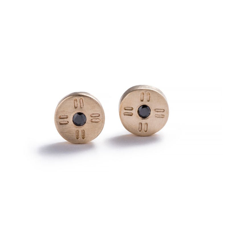 Tiny, circular stud earrings of 14k yellow gold, with subtle engraved details around a round, black diamond. Hand-crafted in Portland, Oregon.