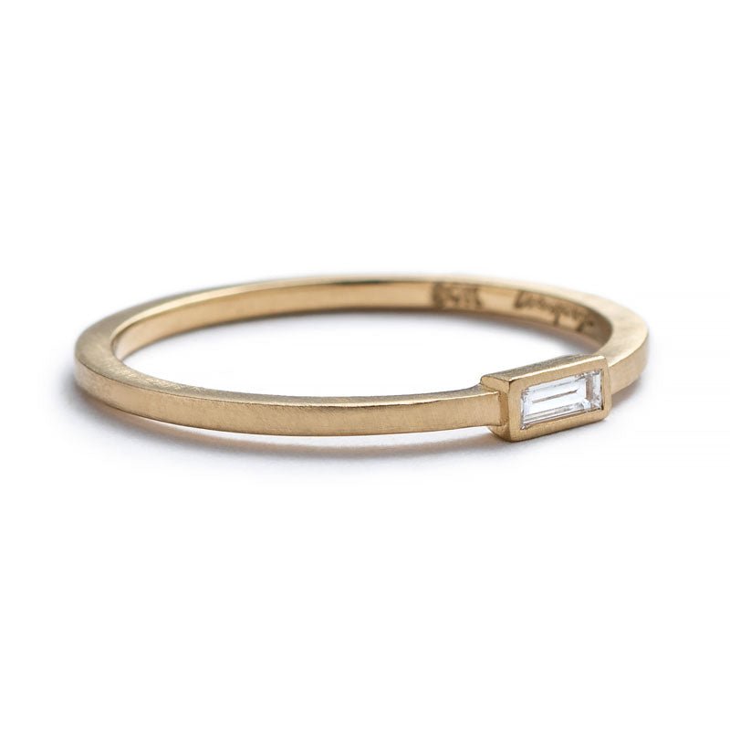 Thin, 14k yellow gold ring with a matte finish, a small, white diamond baguette bezel-set parallel to the band, and the betsy & iya logo engraved inside the band. Hand-crafted in Portland, Oregon.