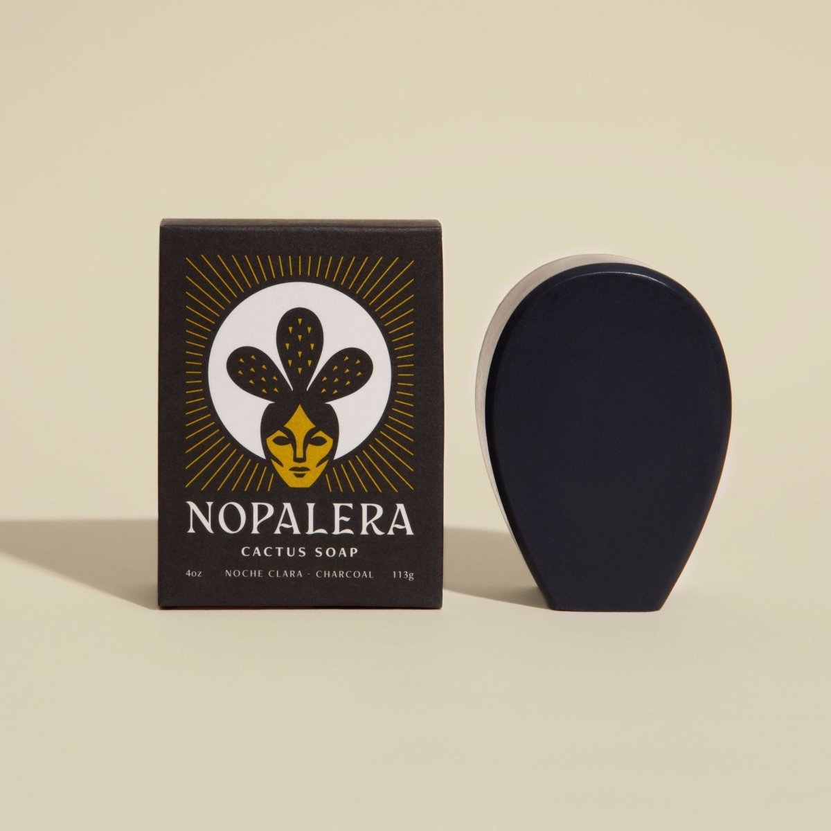 An oval shaped black bar soap stands to the right of its black rectangular packaging. This cactus soap contains a subtle scent of eucalyptus and sage oil with activated charcoal. Noche Clara Cactus Soap is from Nopalera and made in Brooklyn, NY.