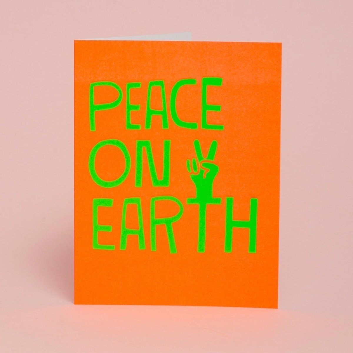 A bright orange card with an illustration of a hand making a peace sign reads: "PEACE ON EARTH." Designed by Ashkahn and printed in Portland, Oregon.