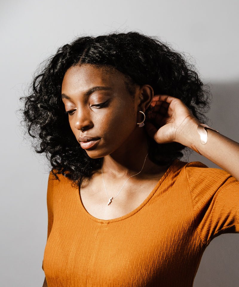 Modern, parentheses-shaped bronze studs with light blue cotton fringe; pictured on a model wearing an ochre dress, a bronze betsy & iya Willamette cuff bracelet, and a bronze betsy & iya Amare necklace.