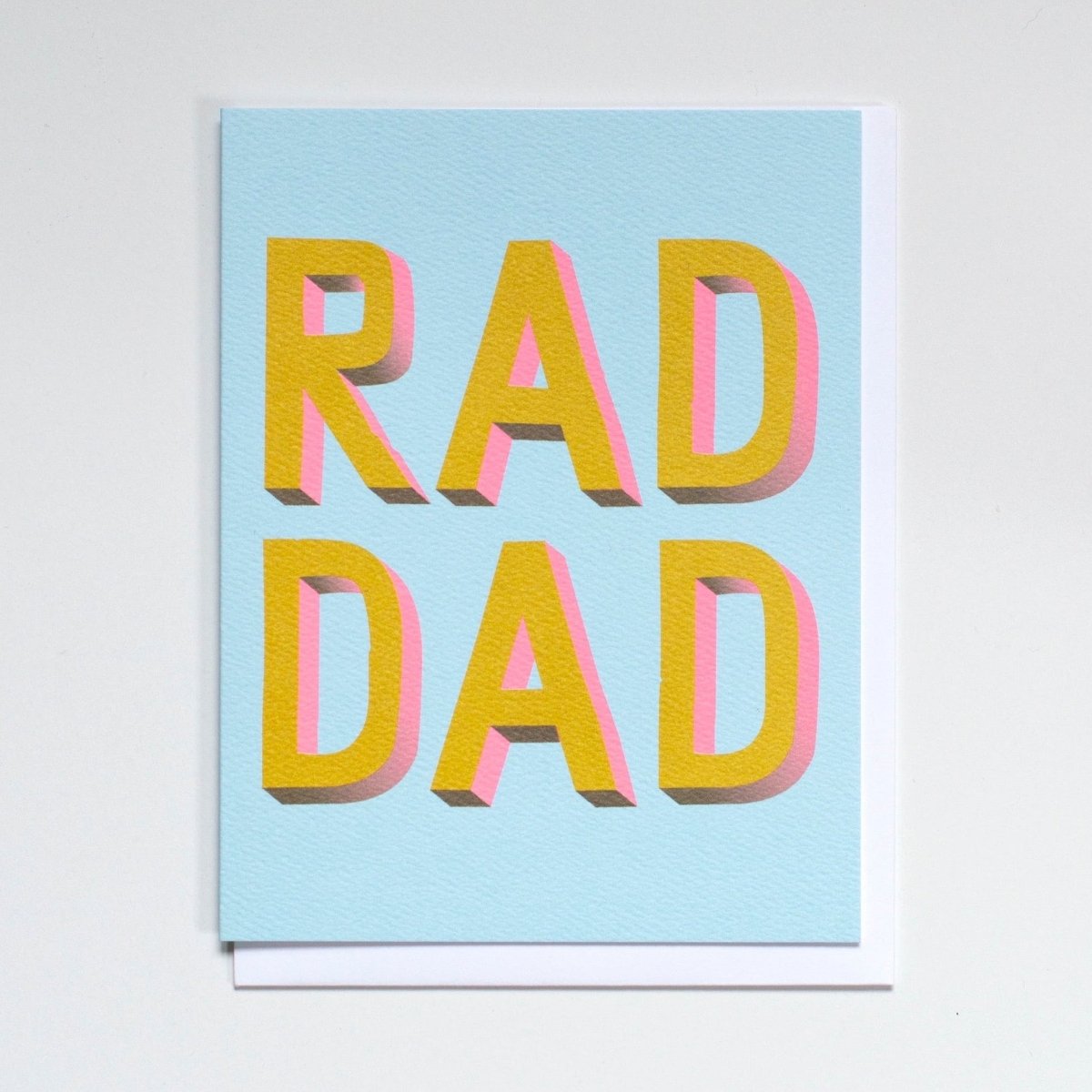 Greeting card reads "RAD DAD" in yellow and pink print against a light blue background. Printed by Banquet Atelier in Vancouver, Canada.