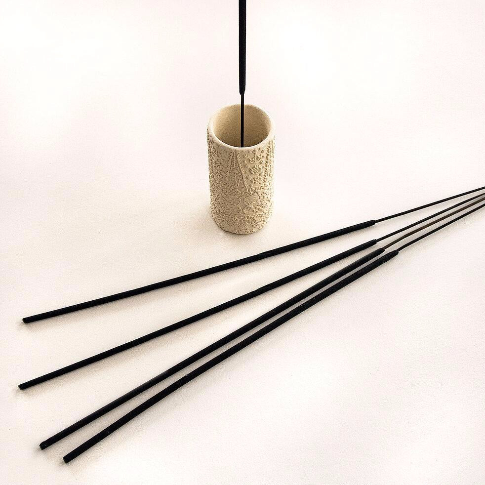 Cream colored cylindrical incense burner with an external embossed pattern. Holds a single stick of black incense. Four sticks of black incense lay in front of holder. Made by Skeem Design in Philadelphia, PA.