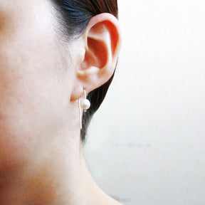 A model wears a gold tone u-shaped earring with a single freshwater pearl. The Short Arc Threader earrings with Large Pearl is designed by Hooks and Luxe and handcrafted in Jackson Heights, NY.