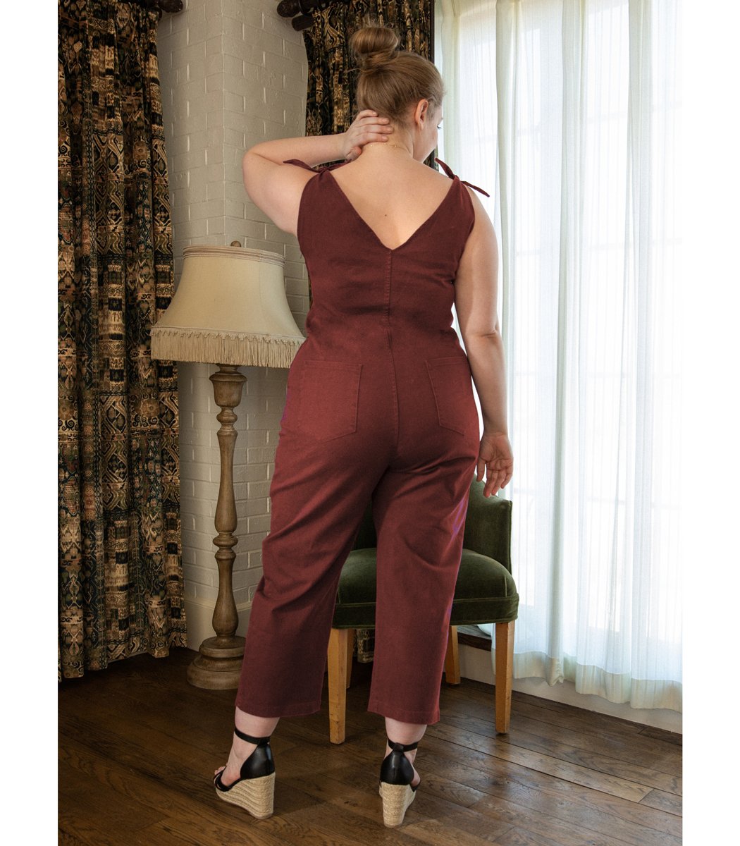 Model shows the back side of a rusty red colored jumpsuit with a V-neckline and tie up shoulder straps. The Slate Overalls in Terracotta are designed by Loup and made in New York City, USA.