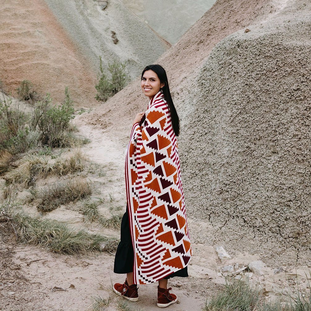 Model wraps a grey, white, gold, chocolate and black pattered throw blanket over their shoulders. The Sunset Mesa Throw is designed by Native American knitwear designer Jennifer Berg and made in New Mexico, USA.