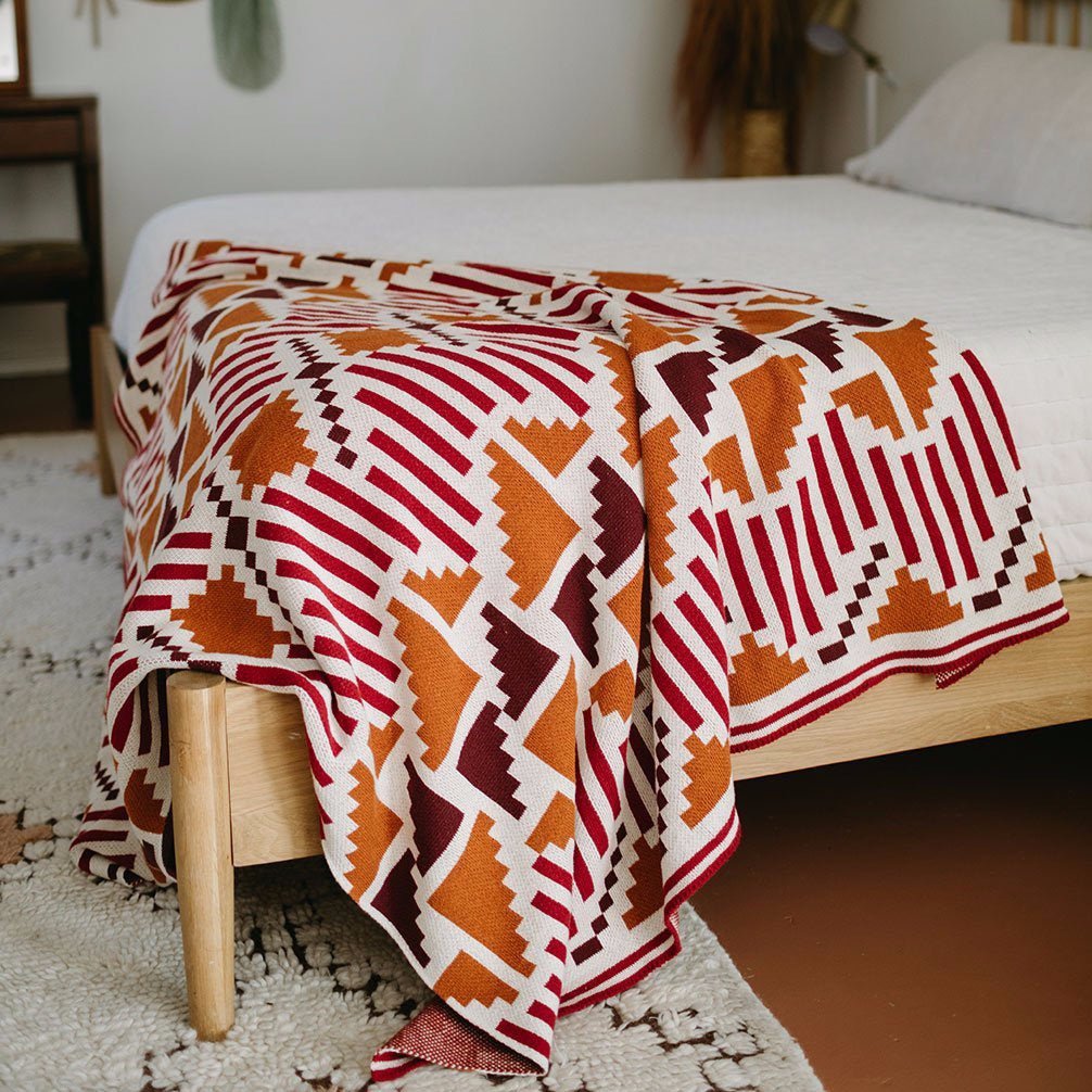 A grey, white, gold, chocolate and black patterned throw displayed on a bed. The Sunset Mesa Throw is designed by Native American knitwear designer Jennifer Berg and made in New Mexico, USA.