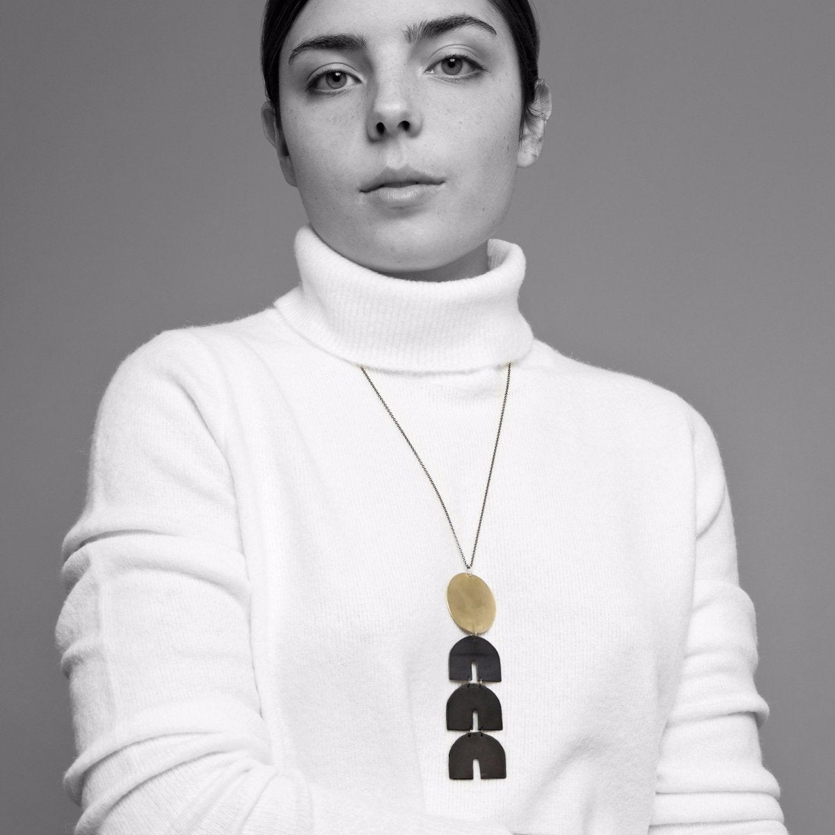 Model wears a four tiered pendant necklace constructed of three oxidized brass arches and one gold tone brass circle. The Telesto necklace is designed and handcrafted by Natalie Joy in Portland, OR.