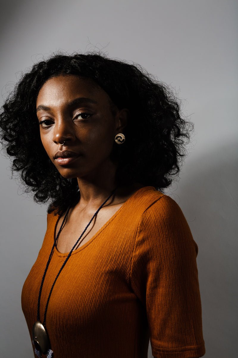 Circular, quarter-sized bronze studs with cutouts and ethically sourced, fair trade lapis lazuli inlays; pictured on a model who wears an ochre dress and a statement betsy & iya Animo Necklace.