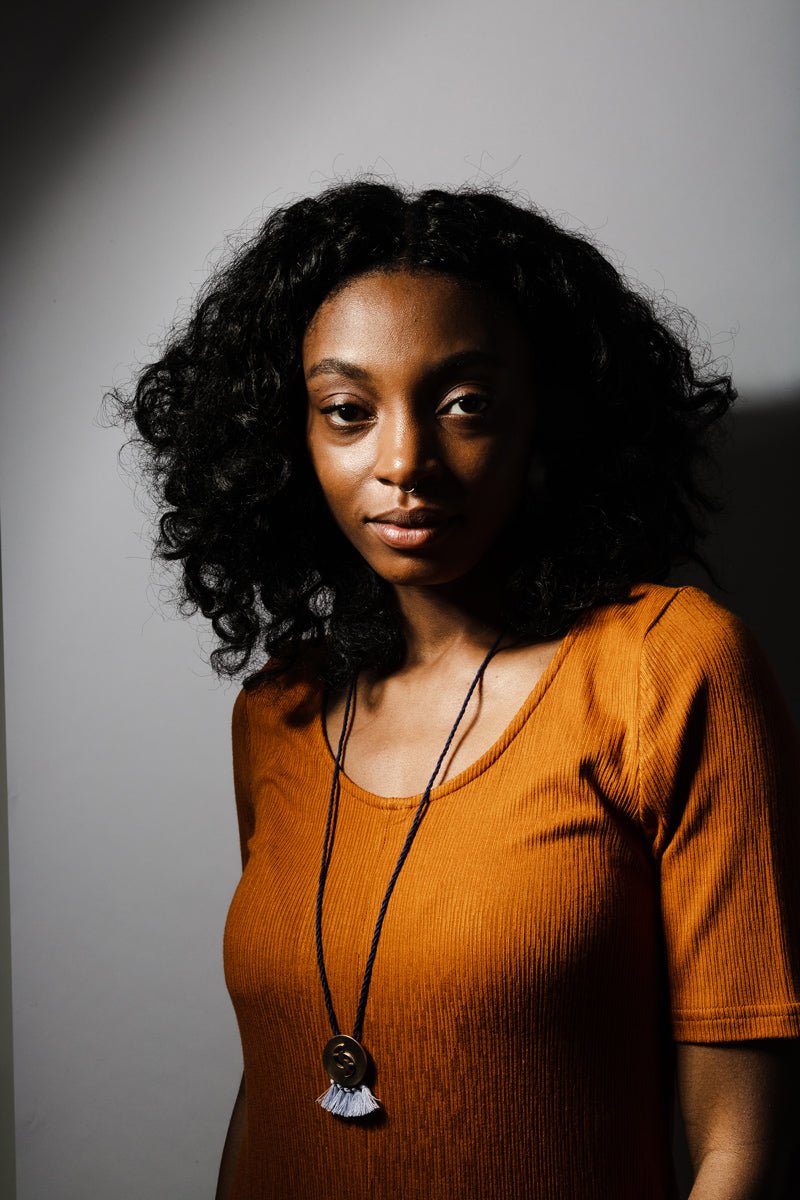The betsy & iya Tenera necklace, with a bronze focal piece, periwinkle fringe, and a navy blue cotton rope, worn long by a model over top of an ochre dress.