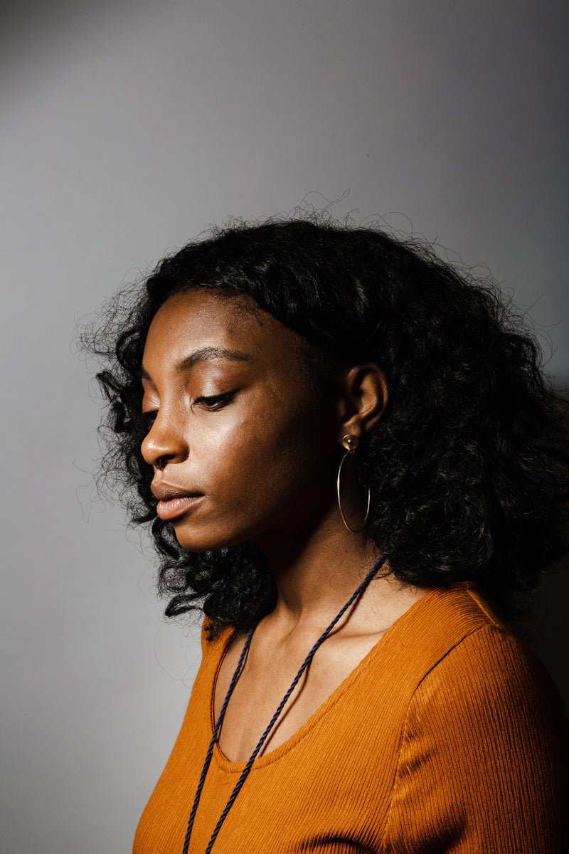 Sleek bronze studs featuring a bezel set, fair trade, and ethically sourced lapis lazuli, with large silver hoops dangling below; pictured on the profile of a model with curly black hair and an ochre dress.