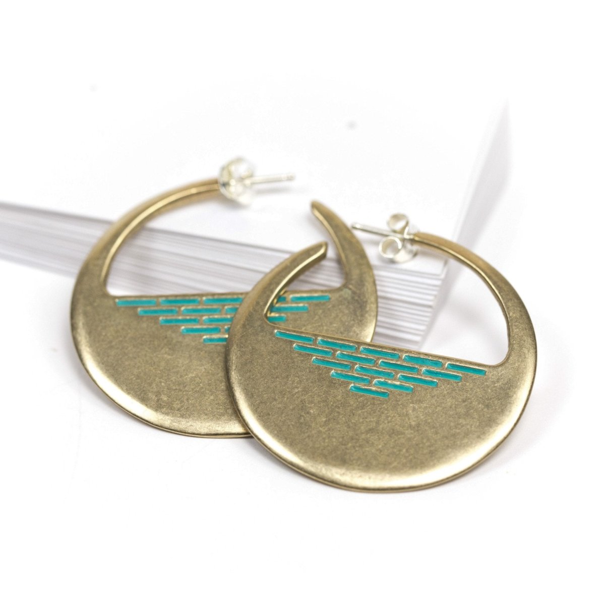 A pair of cast-bronze disc hoop earrings, decorated with a pyramid of geometric, aqua paint detail through the center of the disc, and finished with sterling silver posts and butterfly backings. Hand-crafted in Portland, Oregon.