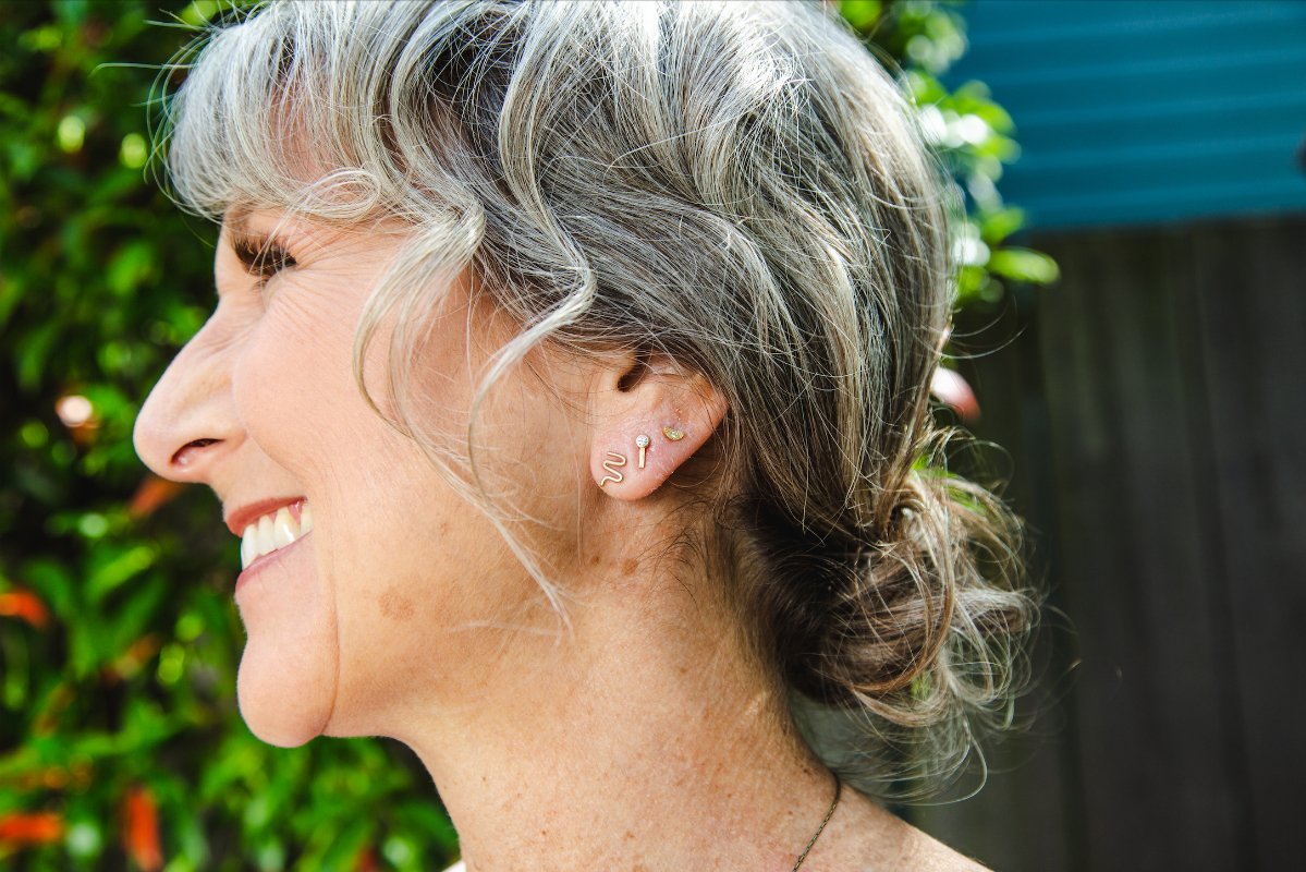 Tiny, half-moon Dulcis stud earrings of 14k yellow gold with round diamond inlays plus two other earring styles worn by model with gray hair.