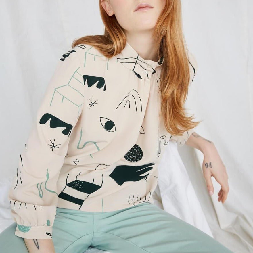 Red-haired woman wearing a cream colored blouse with a black and teal illustrative pattern. The Acacia Top has a buttoned collar and is from Canadian designer Eve Gravel. Illustrative pattern is a collaboration with artist Catherine D'Amours.