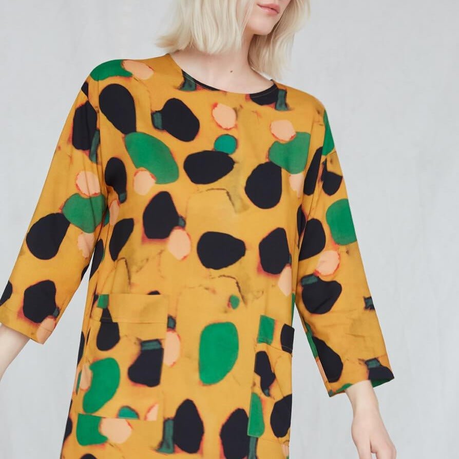 Blonde woman wears a boxy, 3/4 sleeve yellow dress with green, black and pink dots. The Big Trail Dress in Klimt features two large pockets seen in the front. This dress is from Canadian designer Eve Gravel. Illustrative pattern is a collaboration with artist Catherine D'Amours.