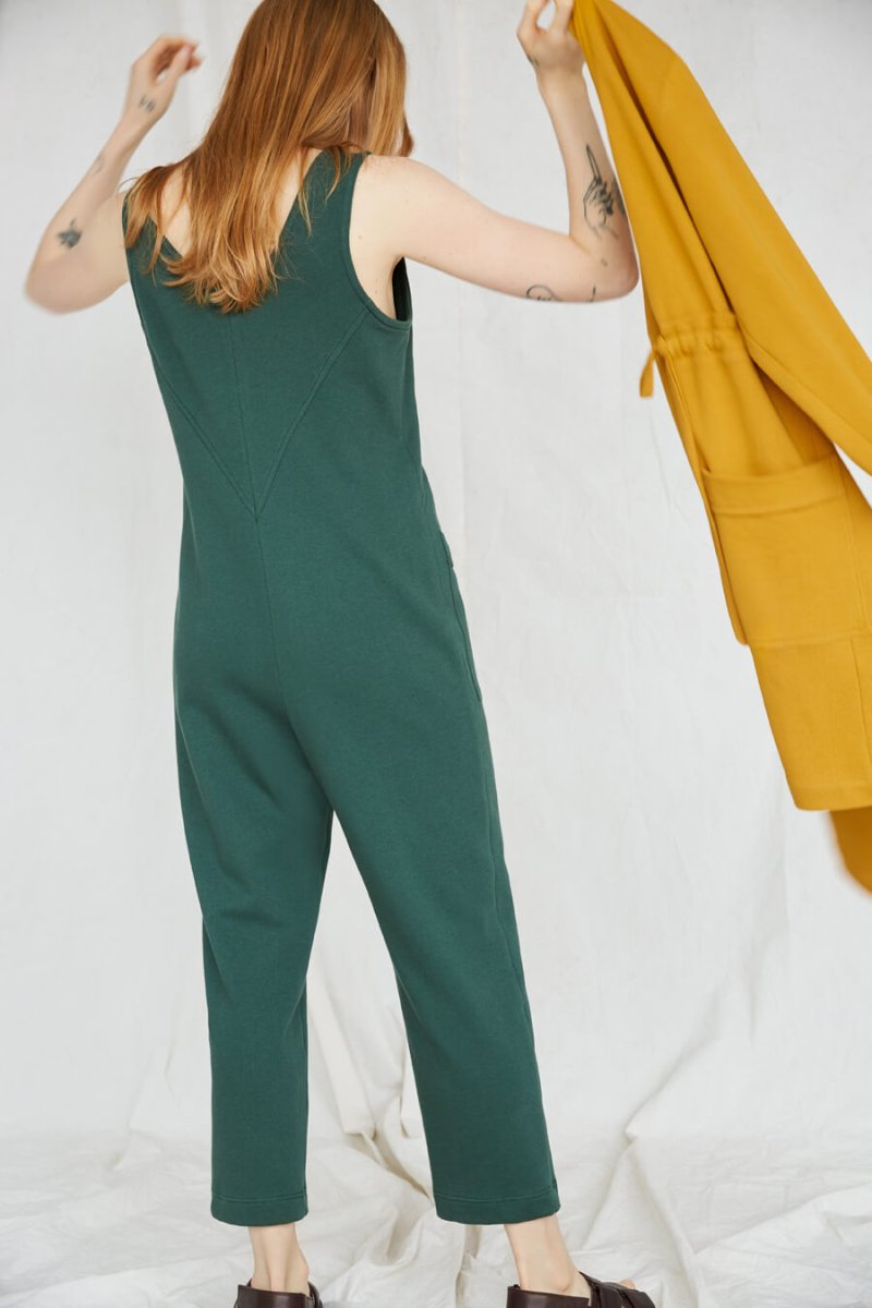 Red-haired woman is facing away and wears an emerald jumpsuit. In her hand is a yellow coat. The Owen Jumpsuit in Spruce is from Canadian designer Eve Gravel.