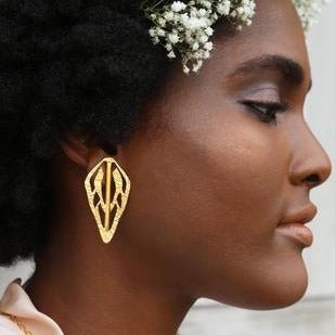 Woman in a profile view wears brass earrings and white flowers in her hair. The Alien Key Post earrings are from designer Lingua Nigra.