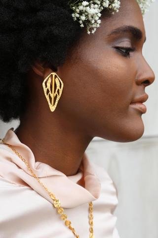 Woman in a profile view wears brass earrings and white flowers in her hair. The Alien Key Post earrings are from designer Lingua Nigra.