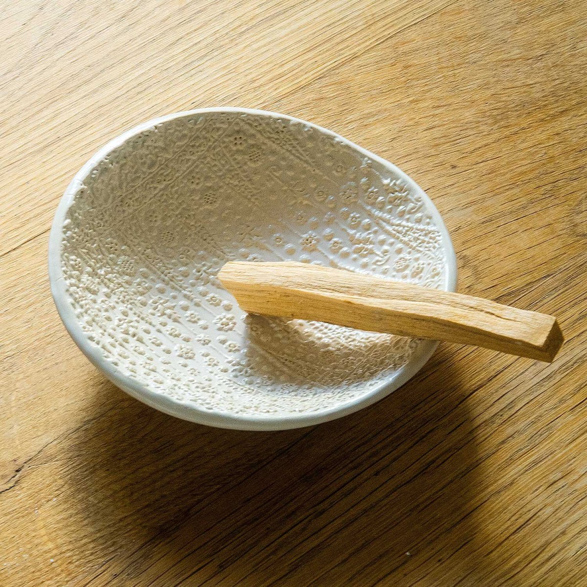 A white embossed offering bowl holding a stick of Palo Santo. Handmade by ceramicist Ellen Hammen.