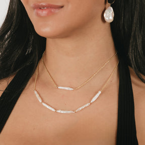 Model wearing two gold necklaces with pearls along with baroque pearl earrings. Top necklace has single pearl bead and bottom necklace has 5 pearl beads.