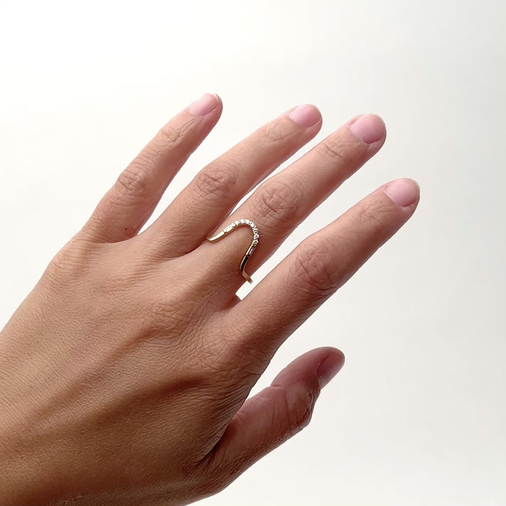 A model wears a narrow curved 14k gold ring on their middle finger. Set in the ring are seven petite round diamonds. Flanking the setting of round diamonds is a baguette white diamond. 
