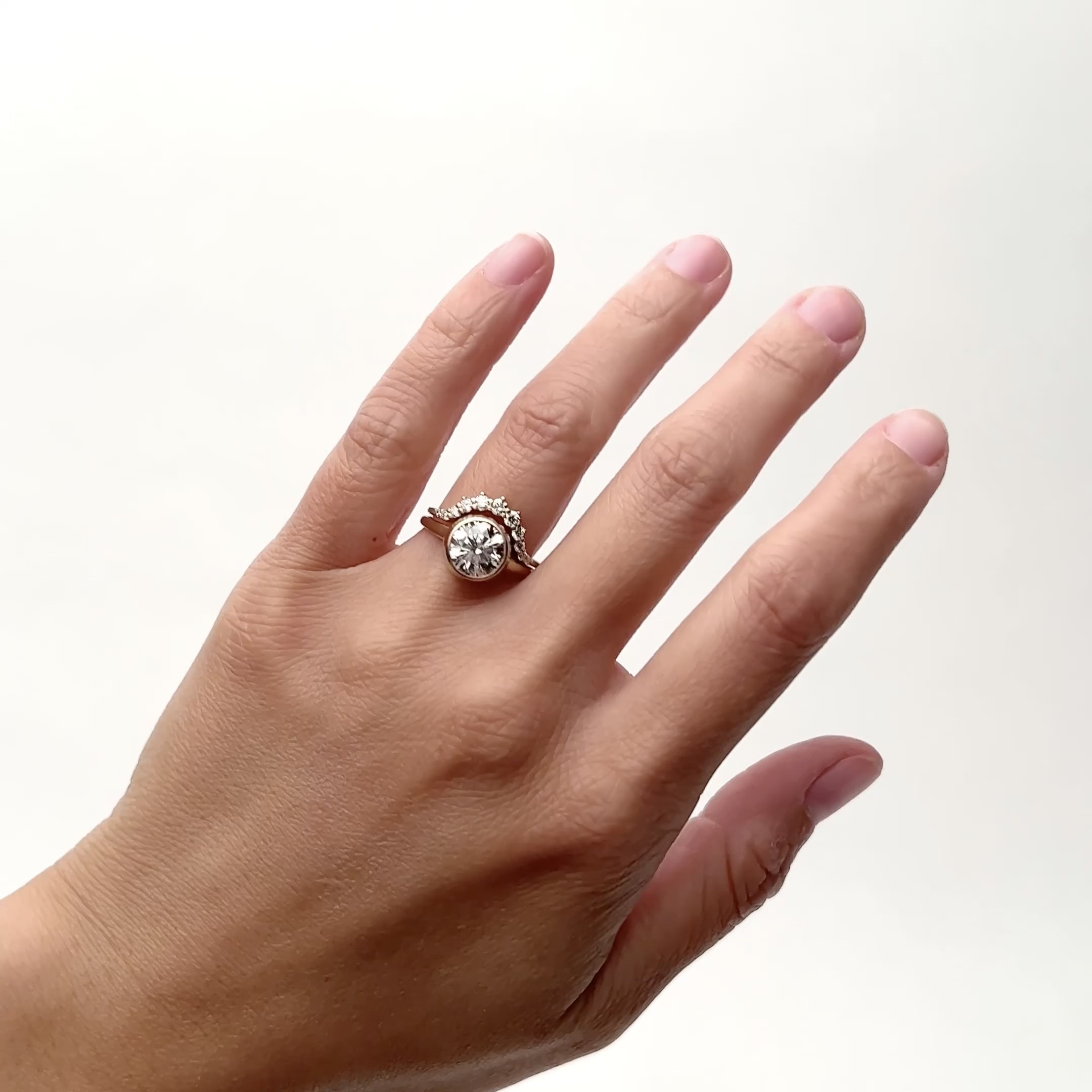 A model wears a rounded narrow band ring with 11 round brilliant cut lab grown diamonds. The model wears the Ortus ring with the 2 carat Salire ring on their left hand ring finger. They rotate their hand from side to side. The Ortus ring and the 2 carat Salire ring is designed and handcrafted in Portland, Oregon.