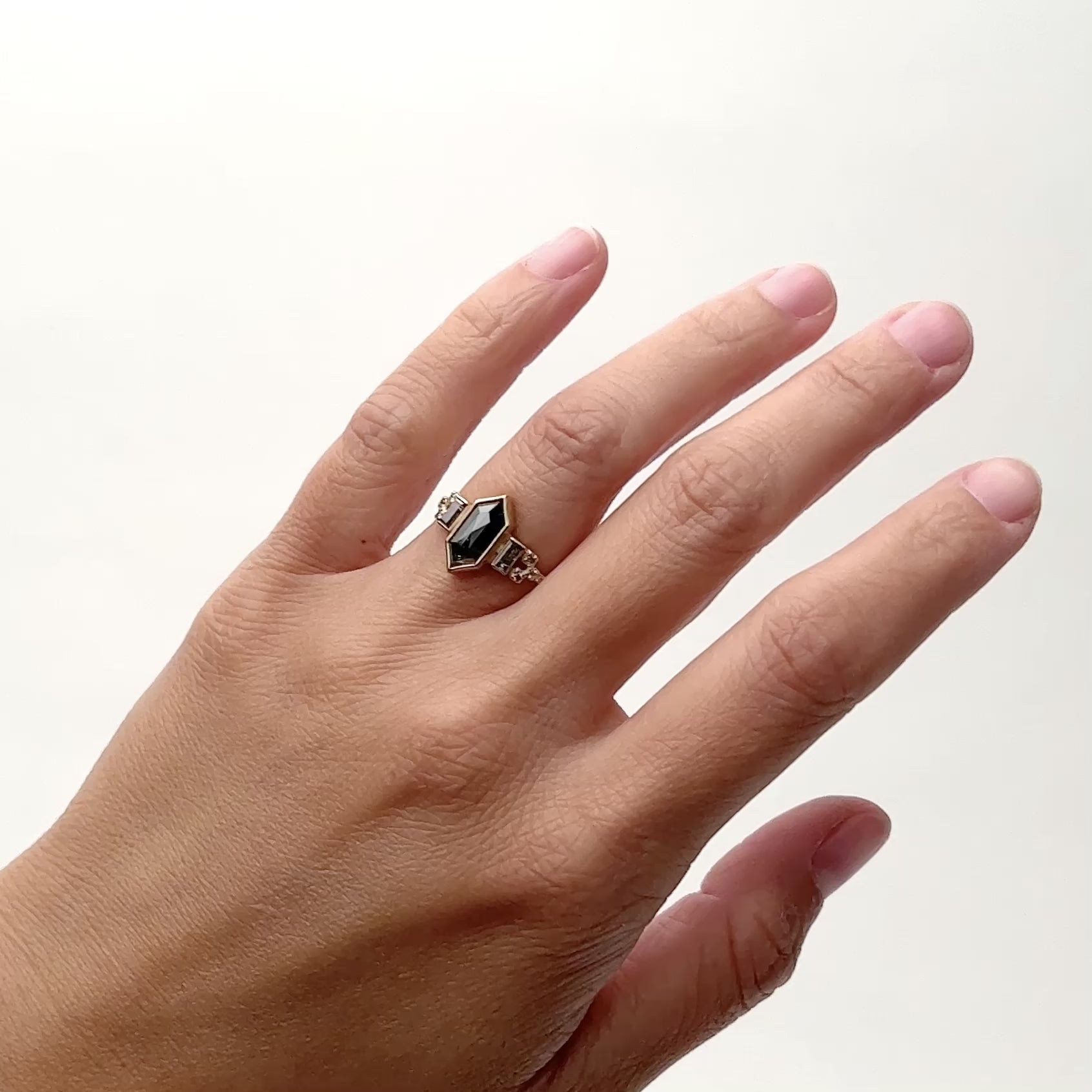 A 14k gold narrow band ring that features a rose cut hexagon salt and pepper diamond. Flanking each side of the diamond are two salt and pepper baguette diamonds. The band forks as it meets the stones. Petite lab grown white diamonds are set in the fork of the band. The Libero ring is designed and handcrafted in Portland, Oregon.