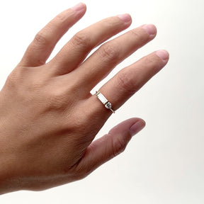 A model wears a narrow band 14k gold ring on their pointer finger. The ring features one large lab grown white baguette diamond and one lab grown round diamond. The baguette diamond lies horizontally along the band and sits high on the finger. The Otium ring is designed and handcrafted in Portland, Oregon.