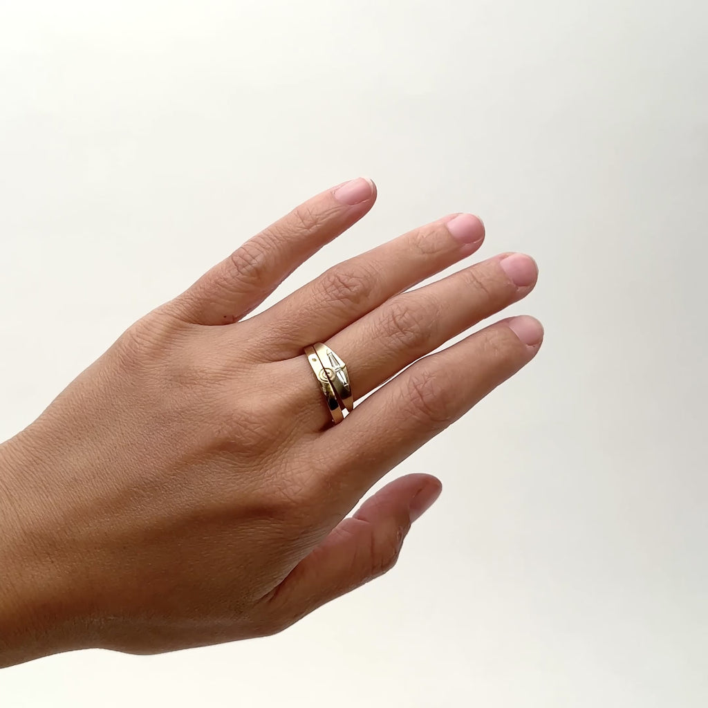 A model wears two 14k yellow gold bands on their middle finger. The south ring features an etched design of half circles (Amandi ring). The north ring widens in the center to form a slight peak (Spero ring). The Spero ring features two white diamond baguettes. Designed and handcrafted in Portland, Oregon.